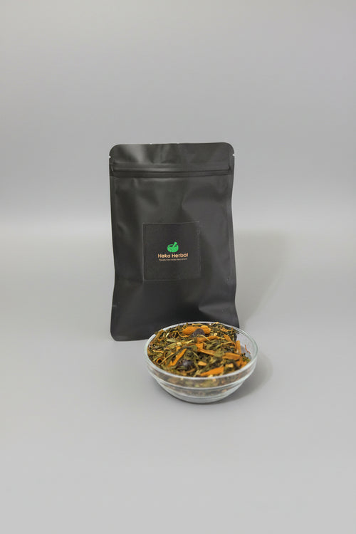 A black stand-up pouch labeled Heka Herbal Metabolic Tea stands on a white marble surface with a small clear bowl of loose tea in front. The background is a dark textured fabric.