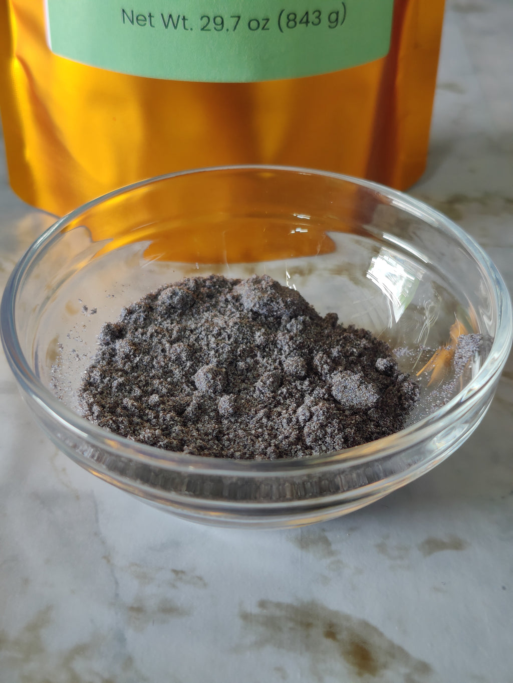 A small clear glass bowl filled with dark gray powder, with an amber-colored pouch and a light green label partially visible in the background on a light marble surface.
