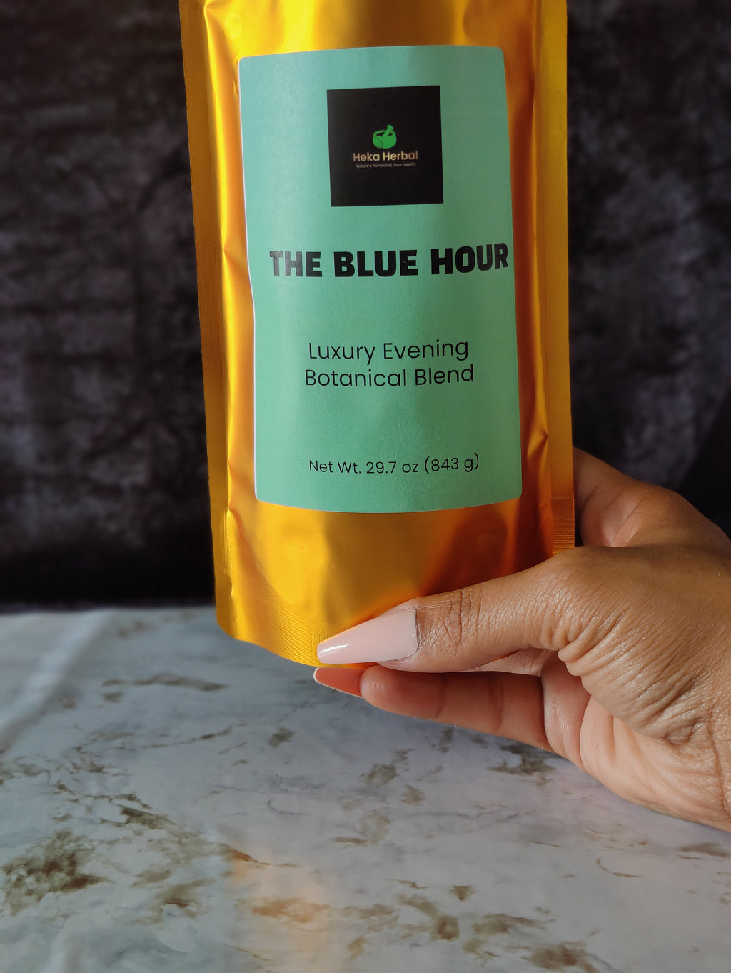 A hand holding a metallic gold stand-up pouch labeled 'THE BLUE HOUR Luxury Evening Botanical Blend' against a dark textured background and a light marble-like surface.